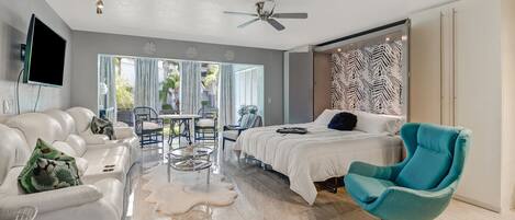 A modern bedroom with a bed, a teal armchair, a white sofa, a glass coffee table, and a ceiling fan.