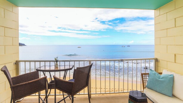 A balcony with wicker chairs and a table, overlooking a beach with boats and a clear sky.