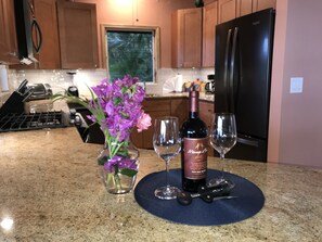 A kitchen counter with a bottle of wine, two glasses, and a vase of purple flowers.