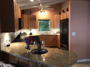 A kitchen with granite countertops, wooden cabinets, a black refrigerator, a window with a view of greenery, and a ceiling fan.