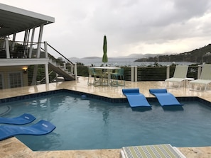 A swimming pool with blue lounge chairs and a table with chairs, overlooking a body of water and distant mountains.