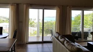 Uma sala de estar ampla com ventilador de teto, sofá, mesa de jantar e grandes portas de vidro deslizantes que dão para uma varanda com vista para o mar.