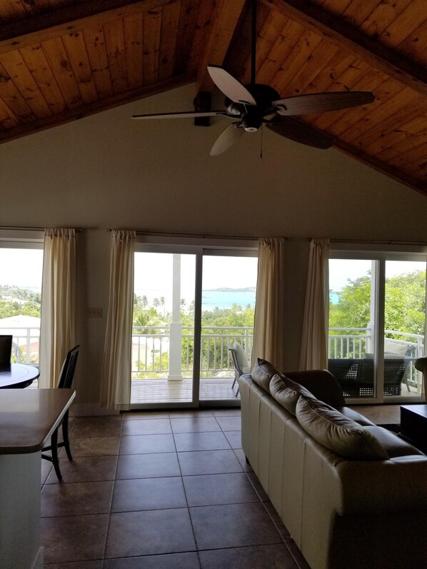 A spacious living area with a ceiling fan, a sofa, a dining table, and large sliding glass doors leading to a balcony with a view of the sea.