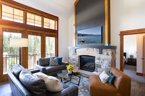 A spacious living room with a stone fireplace, leather sofas, and a large window overlooking a scenic view. - Luxury Top Floor Residence in the Village at Northstar - Catamount 403 (Truckee)