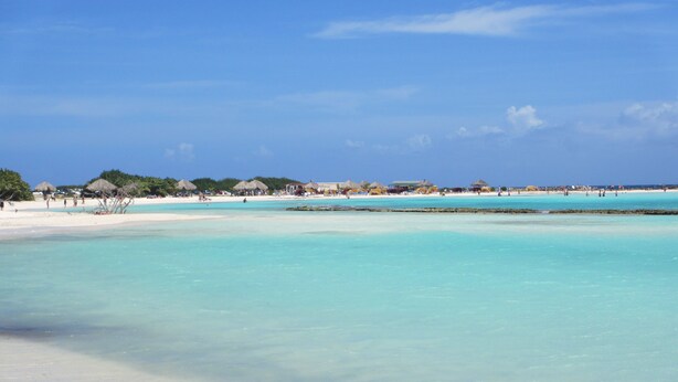 Een strand met helder turquoise water, wit zand en rieten hutten in de verte.