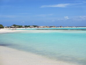 Una playa con aguas turquesas cristalinas, arena blanca y estructuras de techo de paja al fondo.