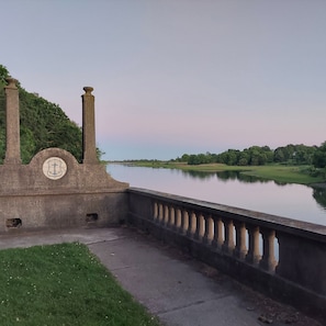 A riverside walkway with a stone monument featuring an anchor symbol, a bridge railing, and a calm river.