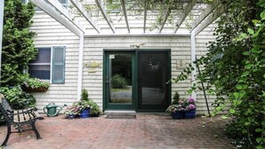 A covered patio with a brick floor, a bench, potted plants, and a door with a welcome sign.