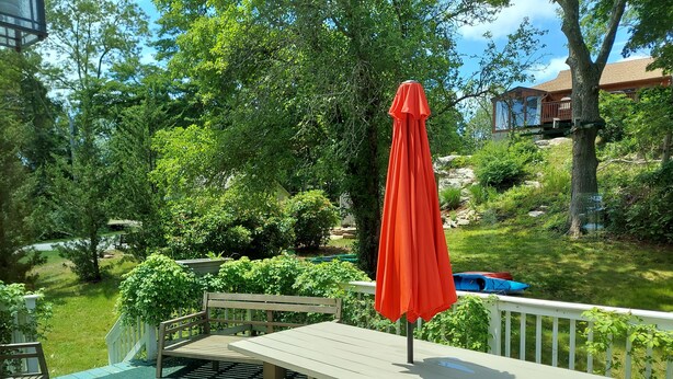 A deck with picnic tables and a red umbrella, surrounded by lush greenery and a house in the background.