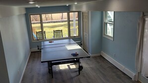 A room with a ping pong table, a window with a view of a lawn and chairs, and a door.