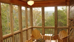 A wooden deck with a table and chairs, large windows offering a forest view, and a ceiling fan.