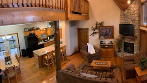 A spacious living area with a stone fireplace, wooden furniture, and a kitchen in the background.