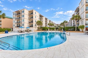A swimming pool with a tiled deck, surrounded by multi-story buildings with balconies, palm trees, and outdoor seating.