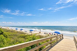 A wooden boardwalk leading to a beach with blue umbrellas and people relaxing.