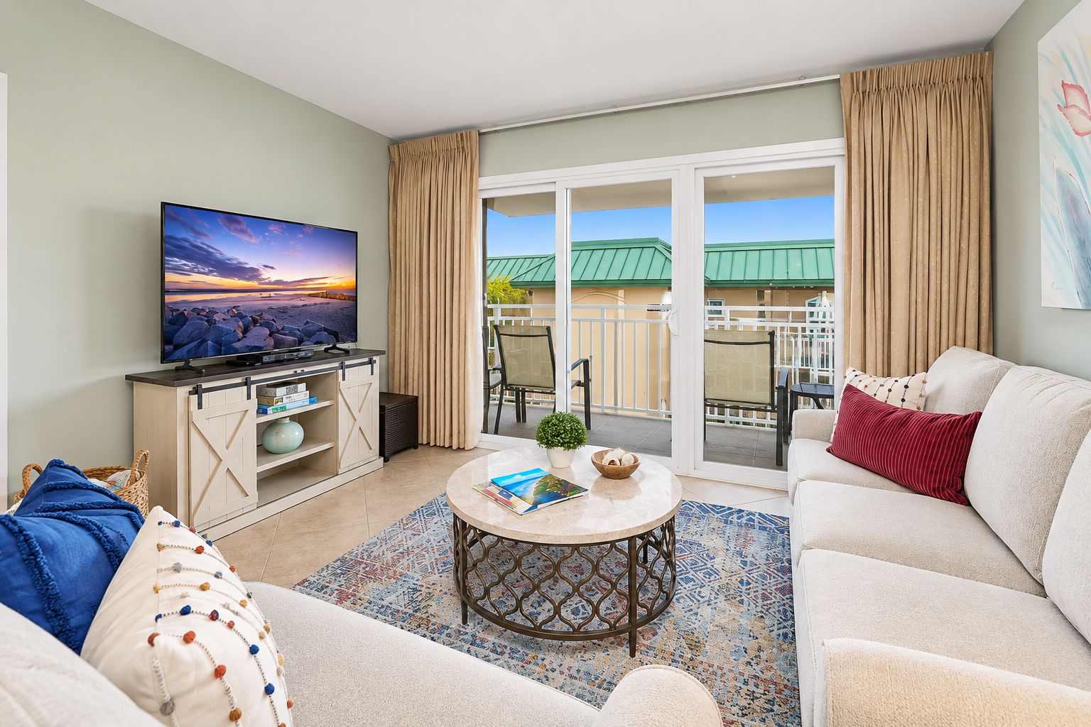 A living room with a television, a sofa, a coffee table, and a view of a balcony with a green roof.