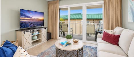 A living room with a television, a sofa, a coffee table, and a view of a balcony with a green roof.