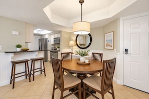 A dining area with a round table, four chairs, a hanging light fixture, and a kitchen in the background.