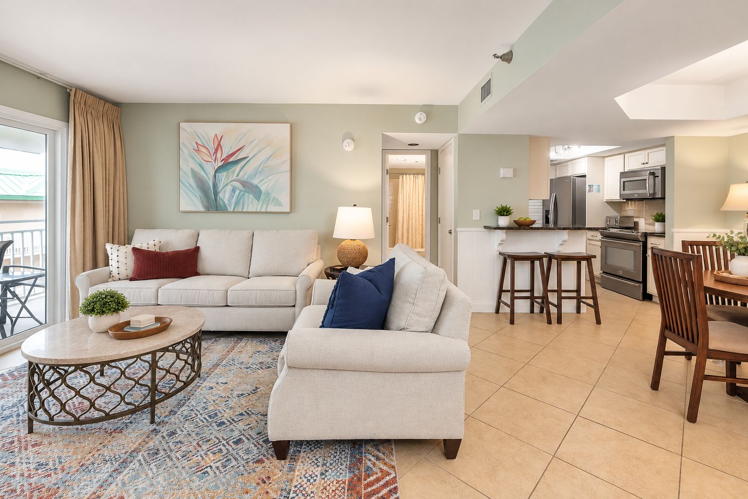 A modern living room with a sofa, coffee table, and dining area. There is a kitchen in the background with appliances and a dining table.