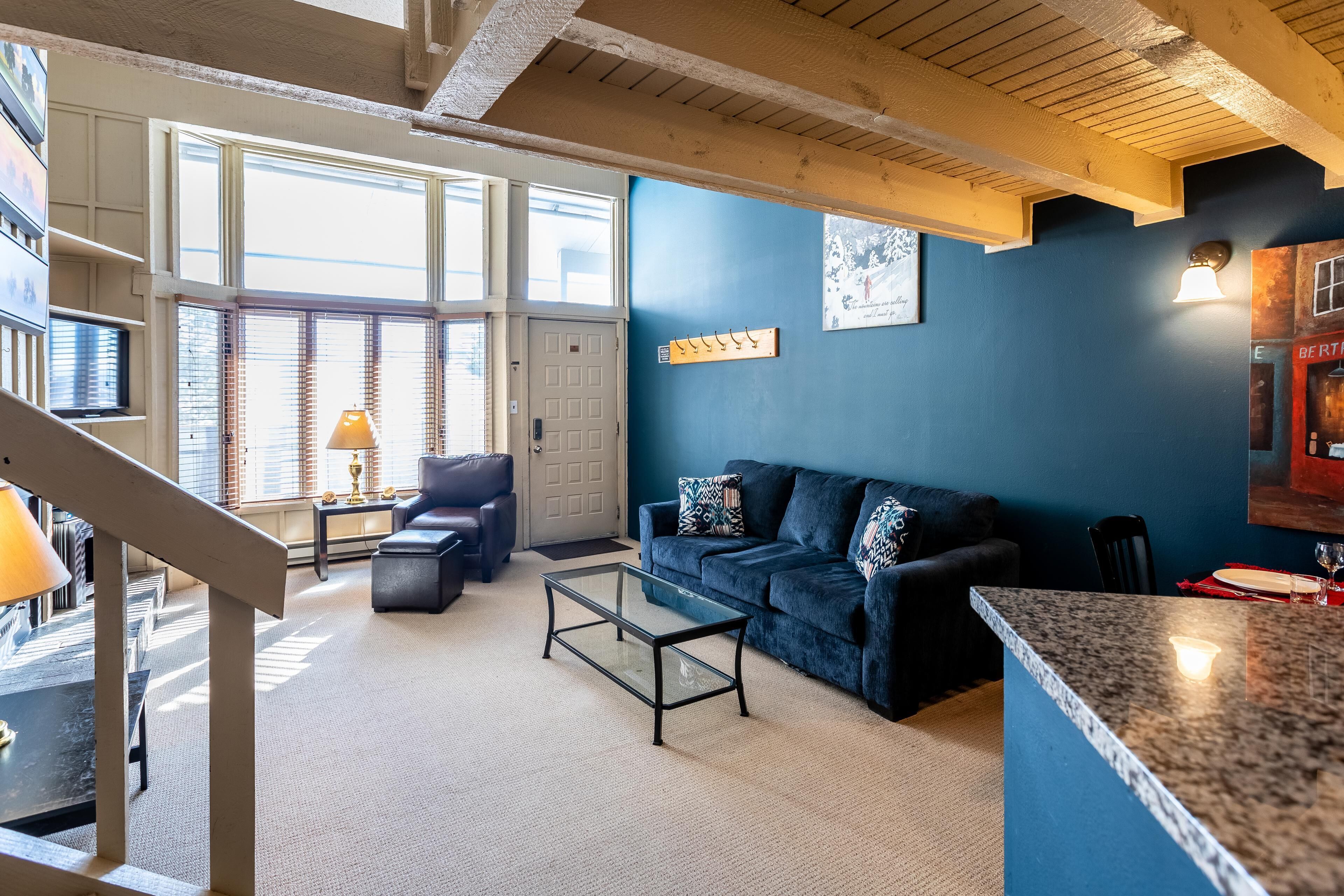 Ein Wohnzimmer mit blauer Wand, einem Glastisch, einem dunkelblauen Sofa und einer Treppe, die zu einer weiteren Ebene führt.