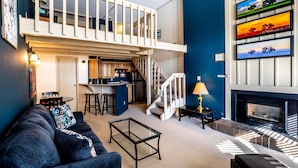 A modern living room with a sofa, coffee table, and a fireplace. A kitchen area is visible in the background, and a staircase leads to an upper level. - ✨ Mountain Village Studio Loft ⎮Fireplace + WIFI + Hot Tubs (Steamboat Springs)