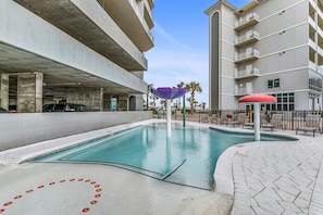 A swimming pool area with a concrete deck, a covered seating area, and a parking garage in the background. - Gulf Front!! Just steps to the Beach- Book Your 5 Star Stay Now!! (Gulf Shores)