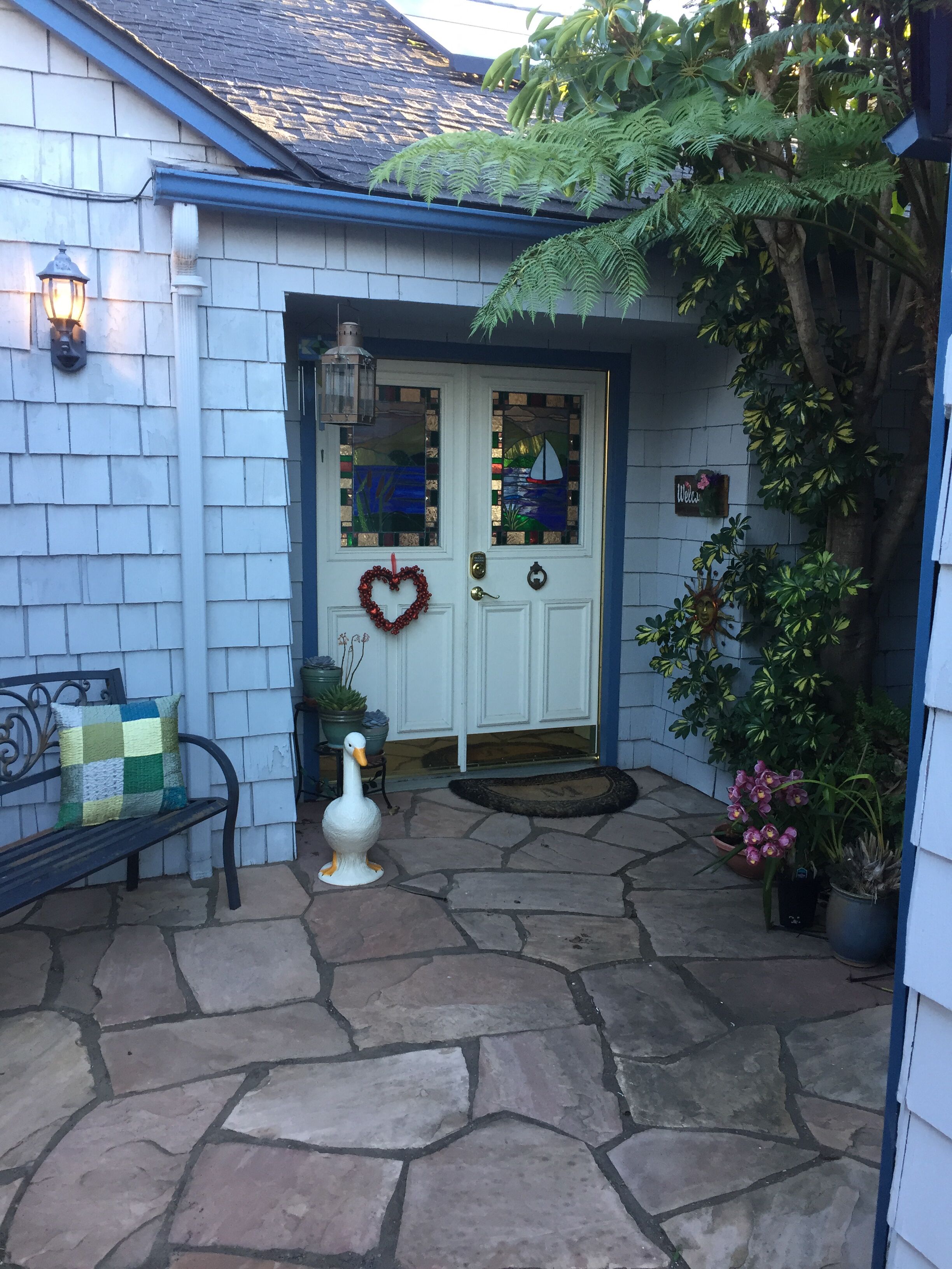 Uma casa com porta branca, um arranjo de grinalda em formato de coração, um enfeite de pato e um banco.