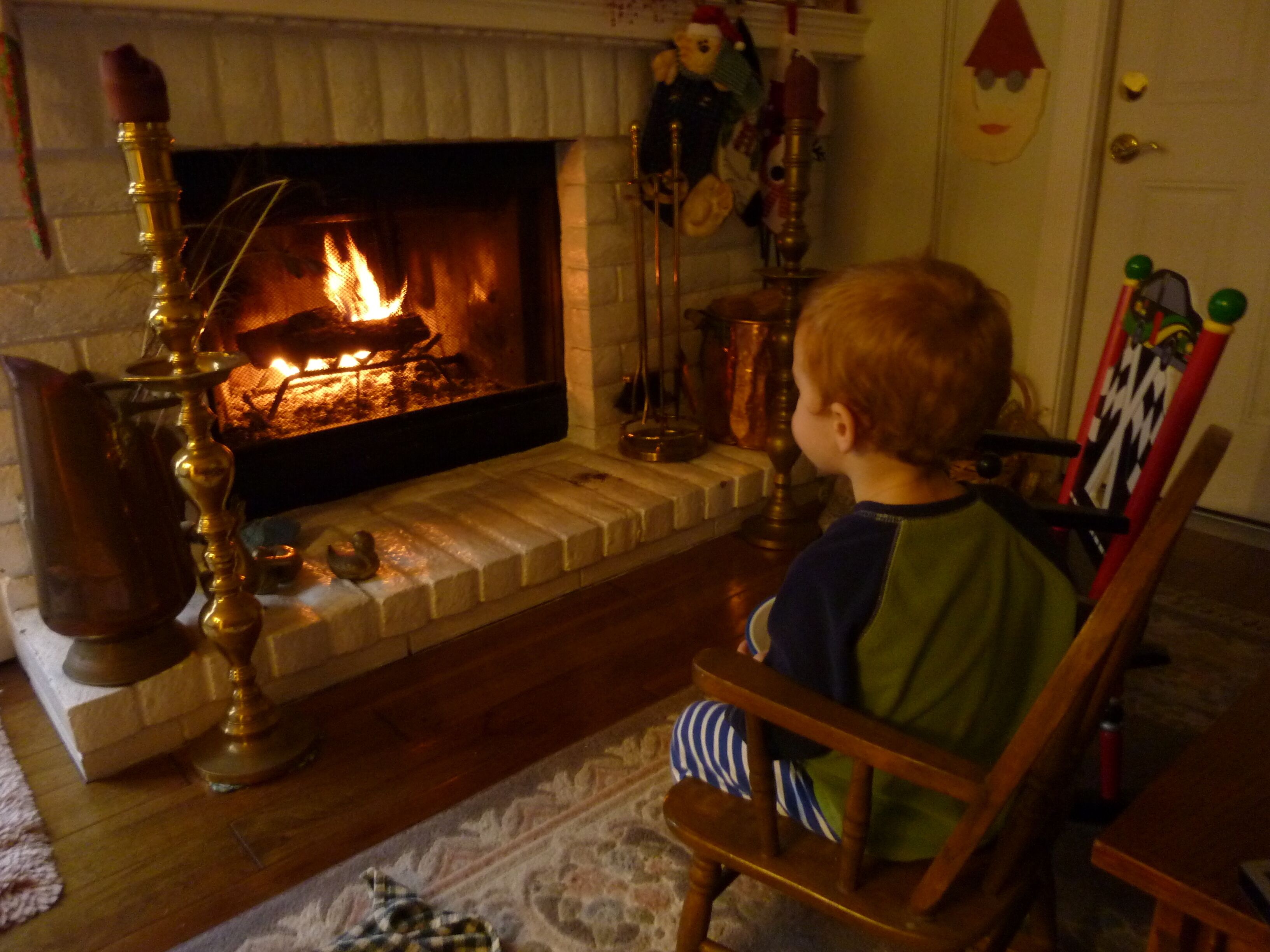 A child sitting on a wooden chair facing a fireplace with a visible fire.