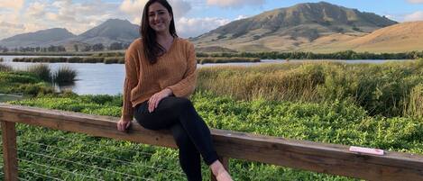 A person sitting on a wooden bench with a view of a lake and hills in the background.