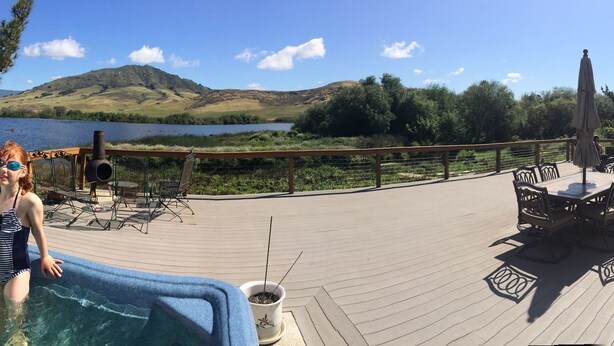 Una terraza con jacuzzi, mobiliario exterior y vistas a un lago y colinas.