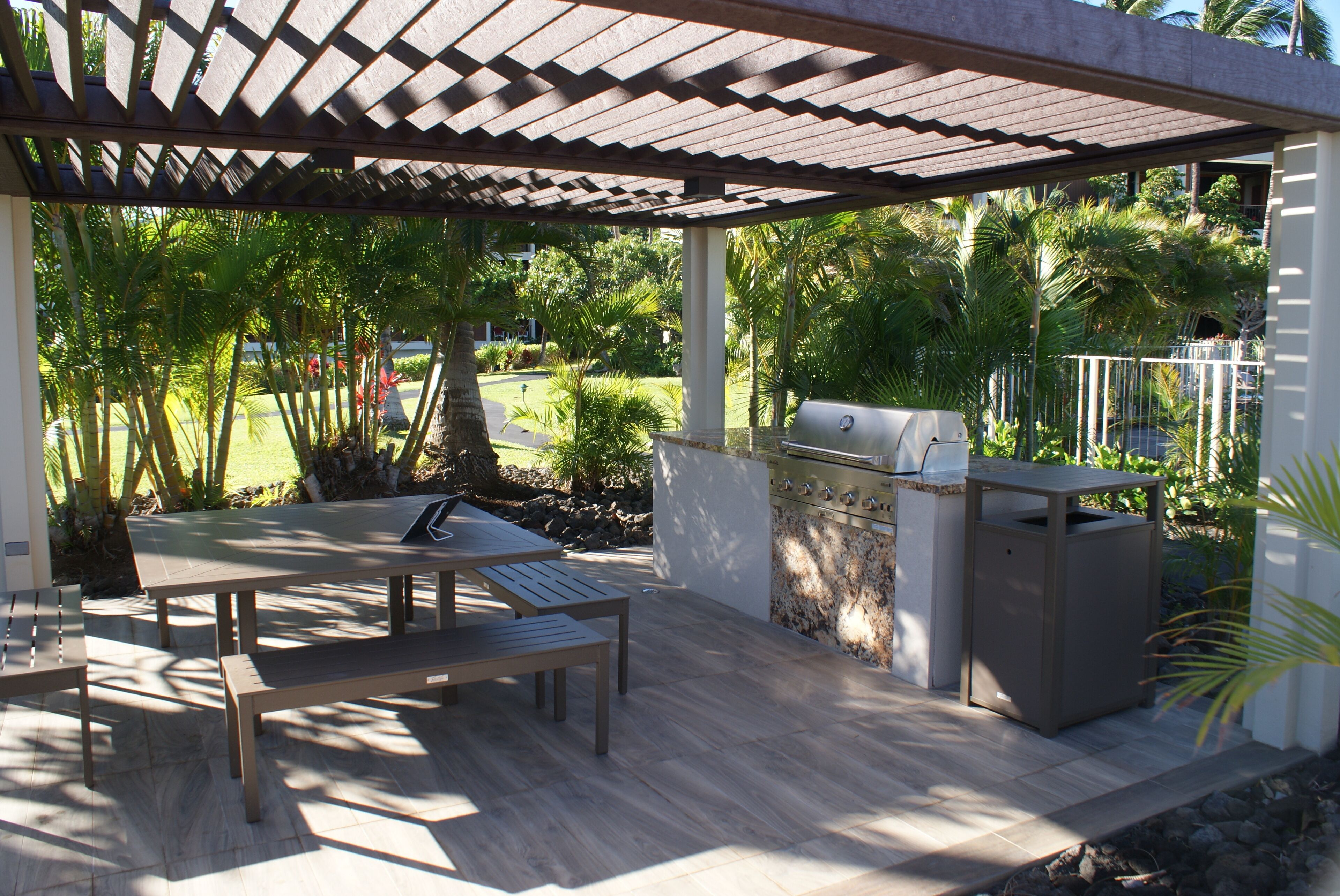 An outdoor patio with a wooden table and bench, a barbecue grill, and a stone wall.