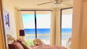 A bedroom with a bed, a ceiling fan, a window with curtains, and a view of the ocean.