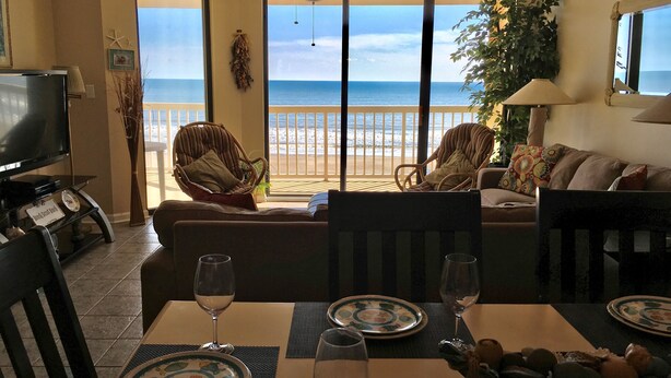 A living room with a view of the beach, a dining table set with plates and glasses, and a television on the left.