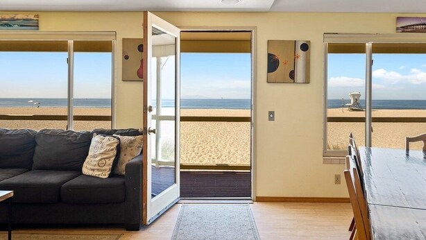 Een kamer met uitzicht op het strand, een zwarte bank, een houten tafel en een deur naar buiten.