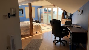 Een slaapkamer met een bed, een bureau met een laptop, een stoel en een raam met uitzicht op een balkon.
