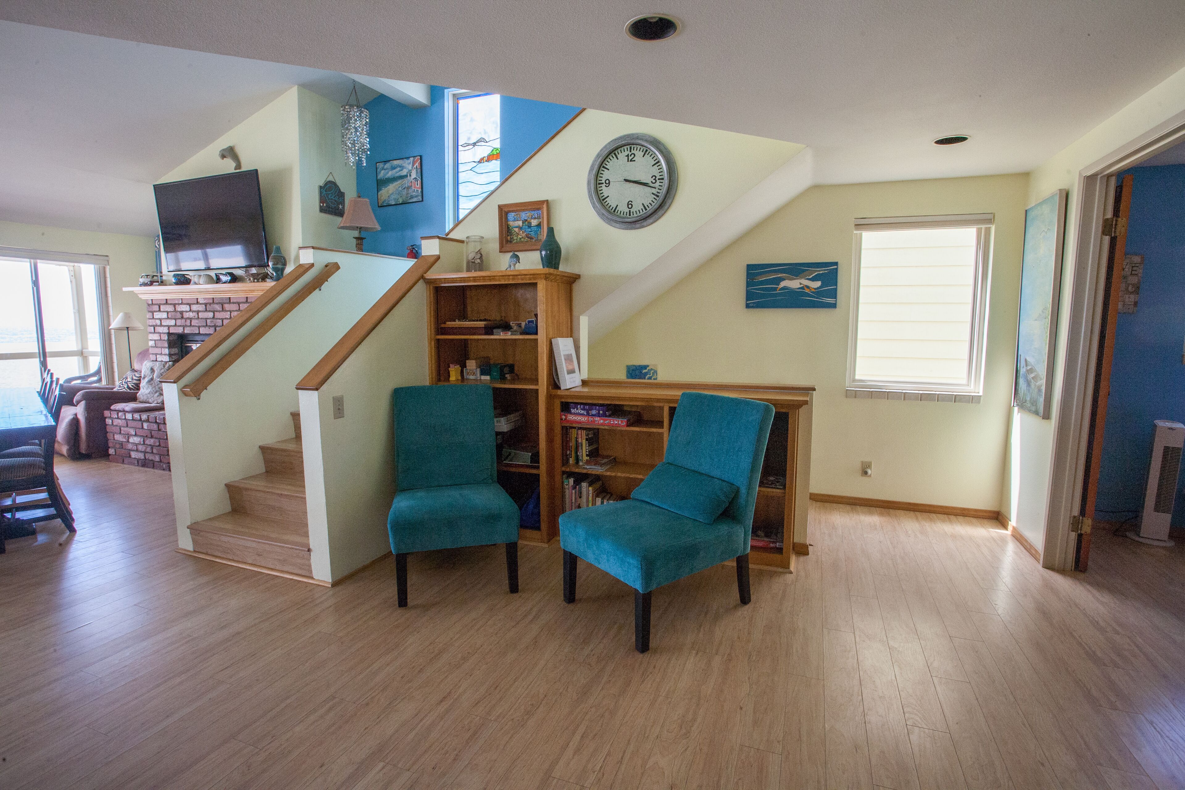 A living room with a staircase, two teal armchairs, a bookshelf, and a clock on the wall.