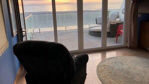 Een kamer met een grote schuifglazen deur die uitkomt op een balkon met uitzicht op zee bij zonsondergang. Er staat een zwarte stoel en er ligt een rond vloerkleed.