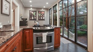 A kitchen with granite countertops, wooden cabinets, a gas stove, and an oven. There are large windows with a view of trees and a deck outside.