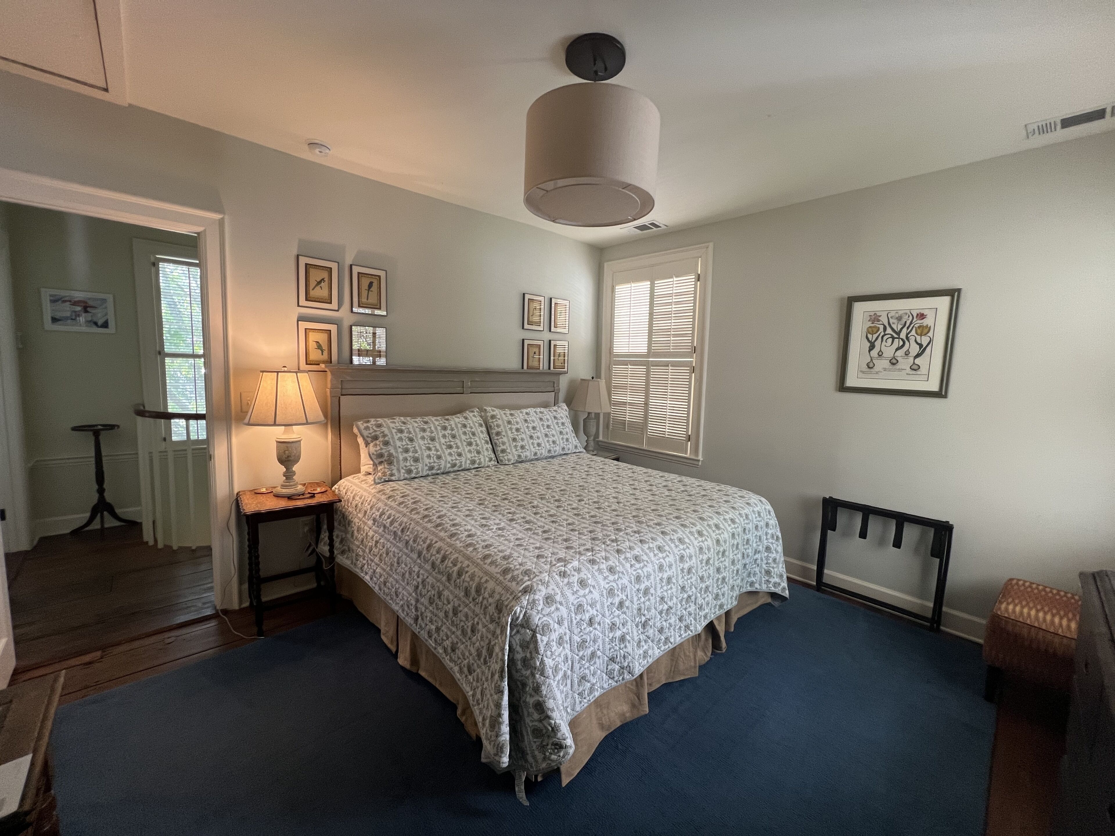 A bedroom with a large bed, a nightstand, a lamp, a window with blinds, and framed pictures on the wall.
