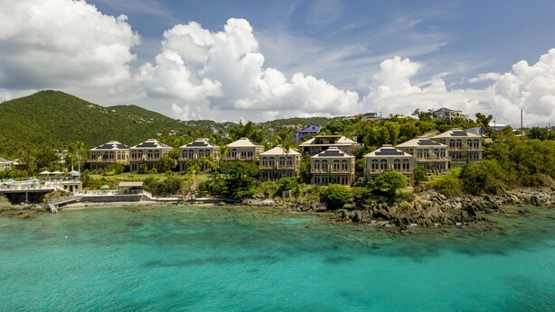 A coastal resort with multiple buildings featuring large windows and balconies, situated near turquoise waters and a mountainous backdrop.