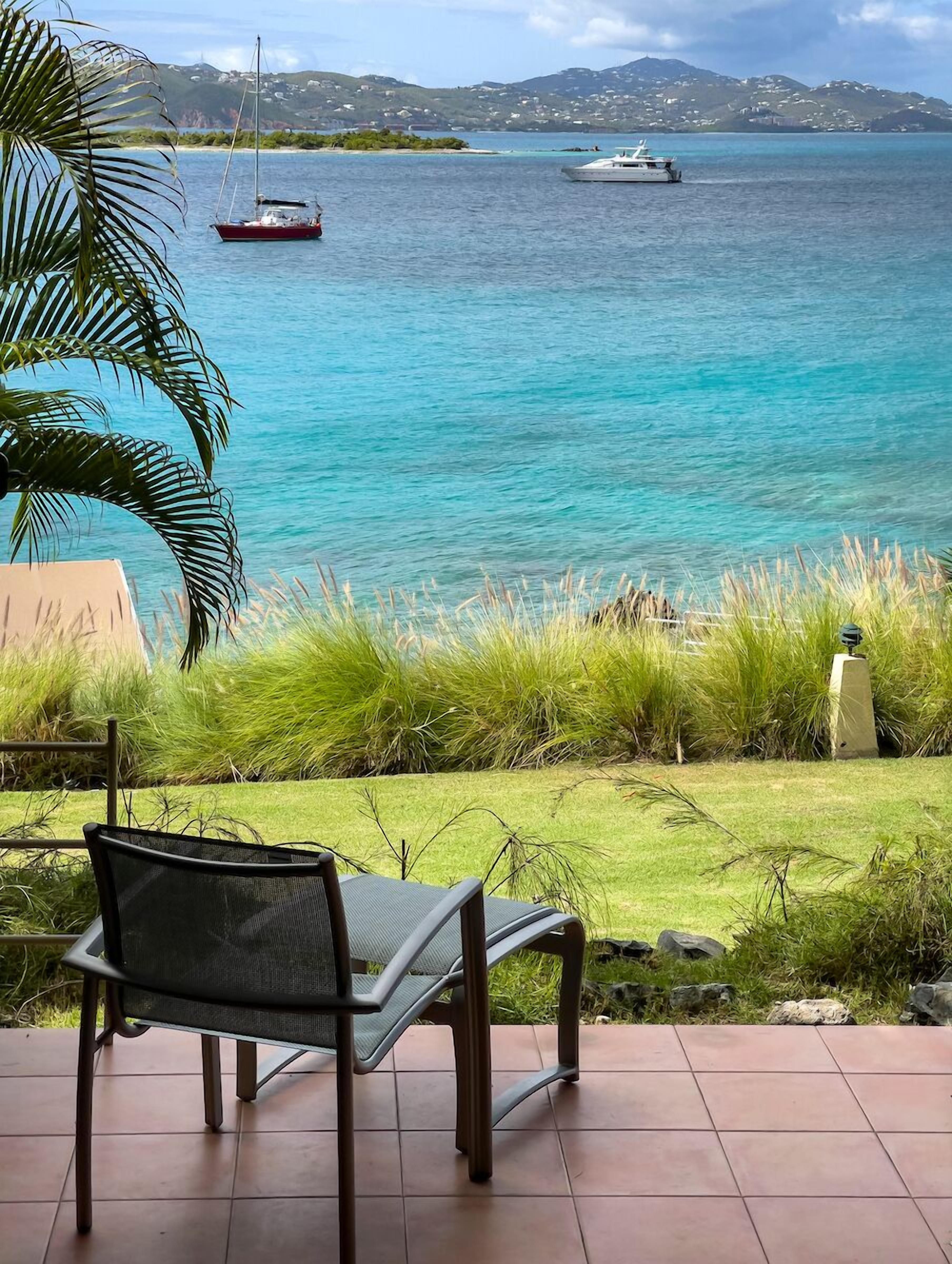 Une terrasse avec une chaise en métal donnant sur une mer turquoise où l’on aperçoit des bateaux, une zone herbeuse et des collines au loin.