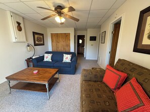 A living room with a ceiling fan, a wooden coffee table, a blue sofa with floral cushions, a brown sofa with patterned cushions, and a framed picture on the wall.