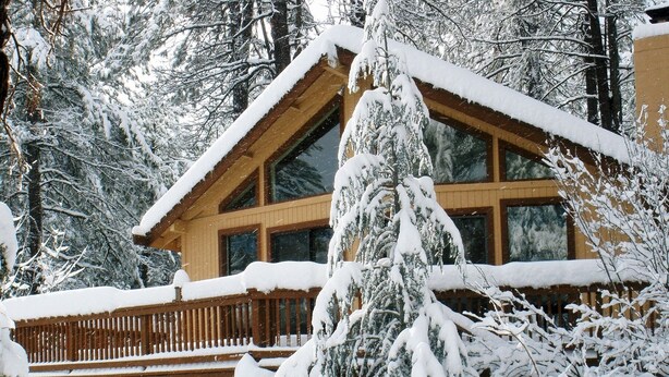 Une cabane en rondins dotée d’une terrasse en bois et de grandes fenêtres, entourée d’arbres couverts de neige.