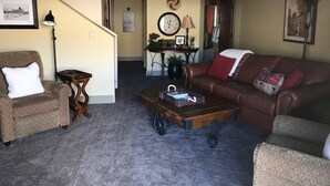 Ein Wohnzimmer mit braunem Ledersofa, einem braunen Sessel, einem Holztisch und einem kleinen Beistelltisch. An der Wand hängt eine Uhr und über dem Sofa ein gerahmtes Bild.