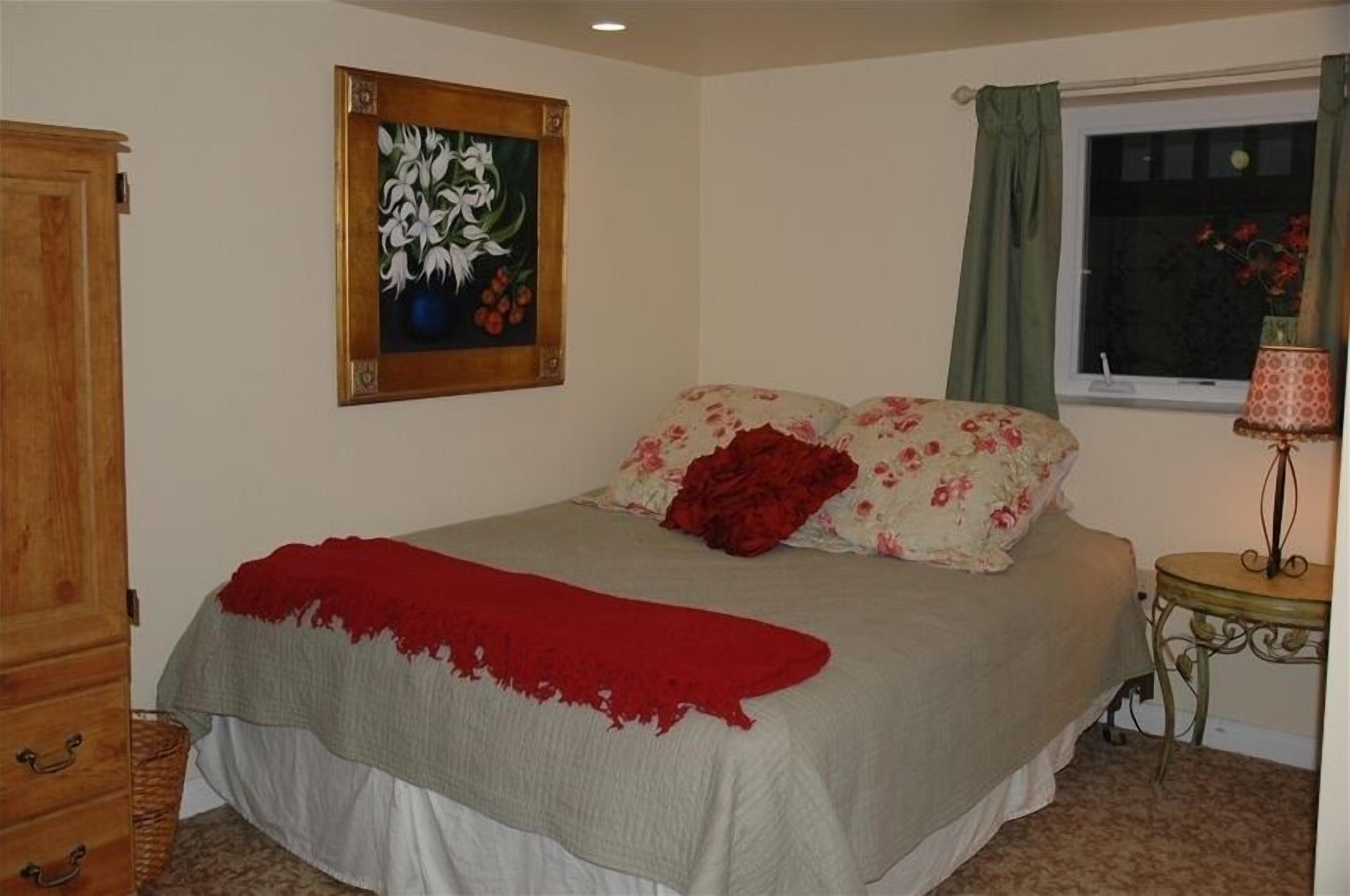 A bedroom with a bed, a wooden wardrobe, a painting, a window with curtains, and a small table with a lamp.