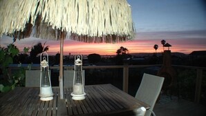 Et træbord med to lygter og en stråtækt parasol på en balkon med udsigt til solnedgang.