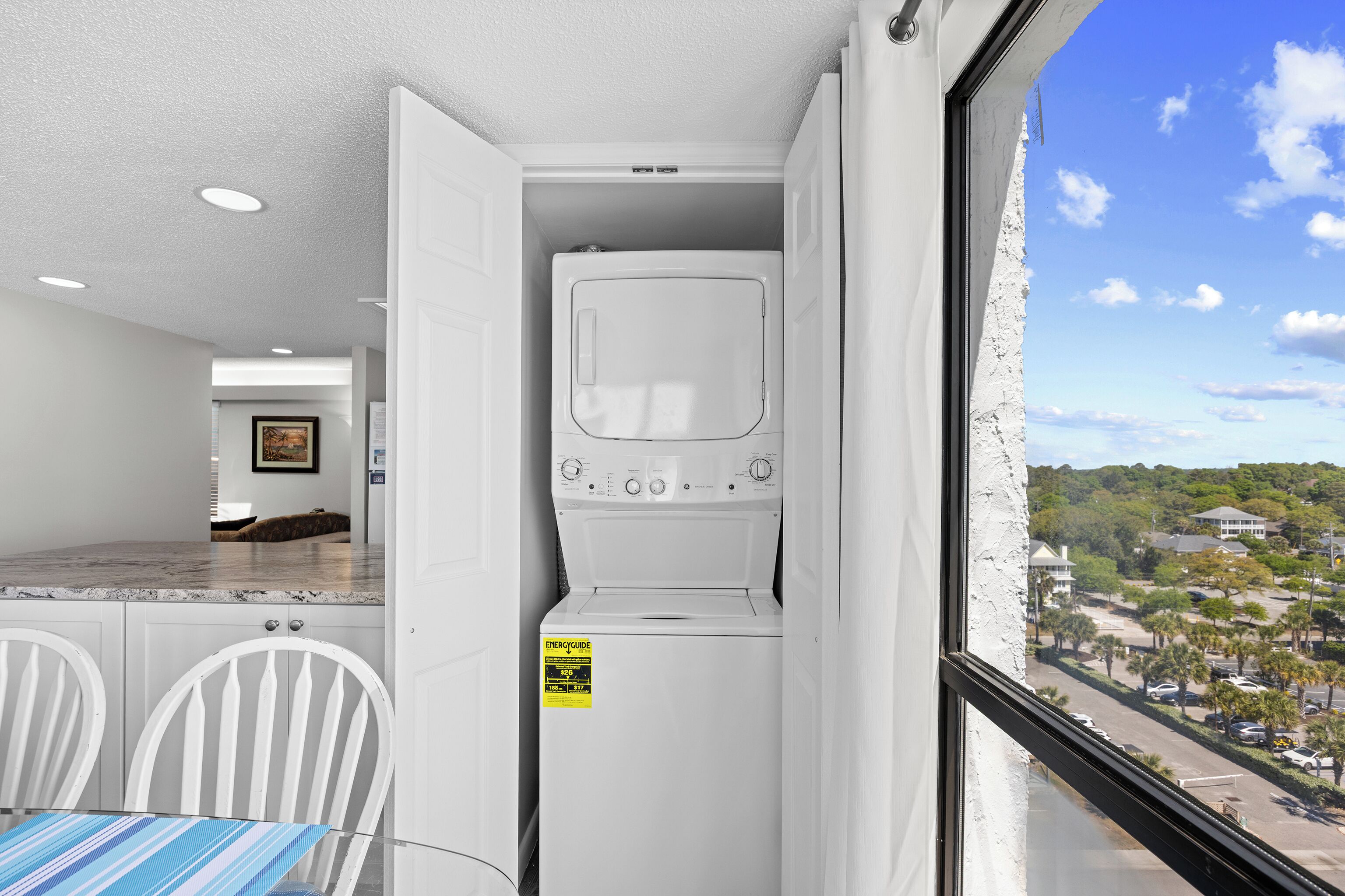A compact laundry room with a washer and dryer, a dining area with a table and chairs, and a view of a residential neighborhood through a window.