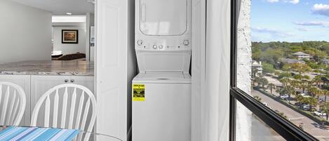 A compact laundry room with a washer and dryer, a dining area with a table and chairs, and a view of a residential neighborhood through a window.