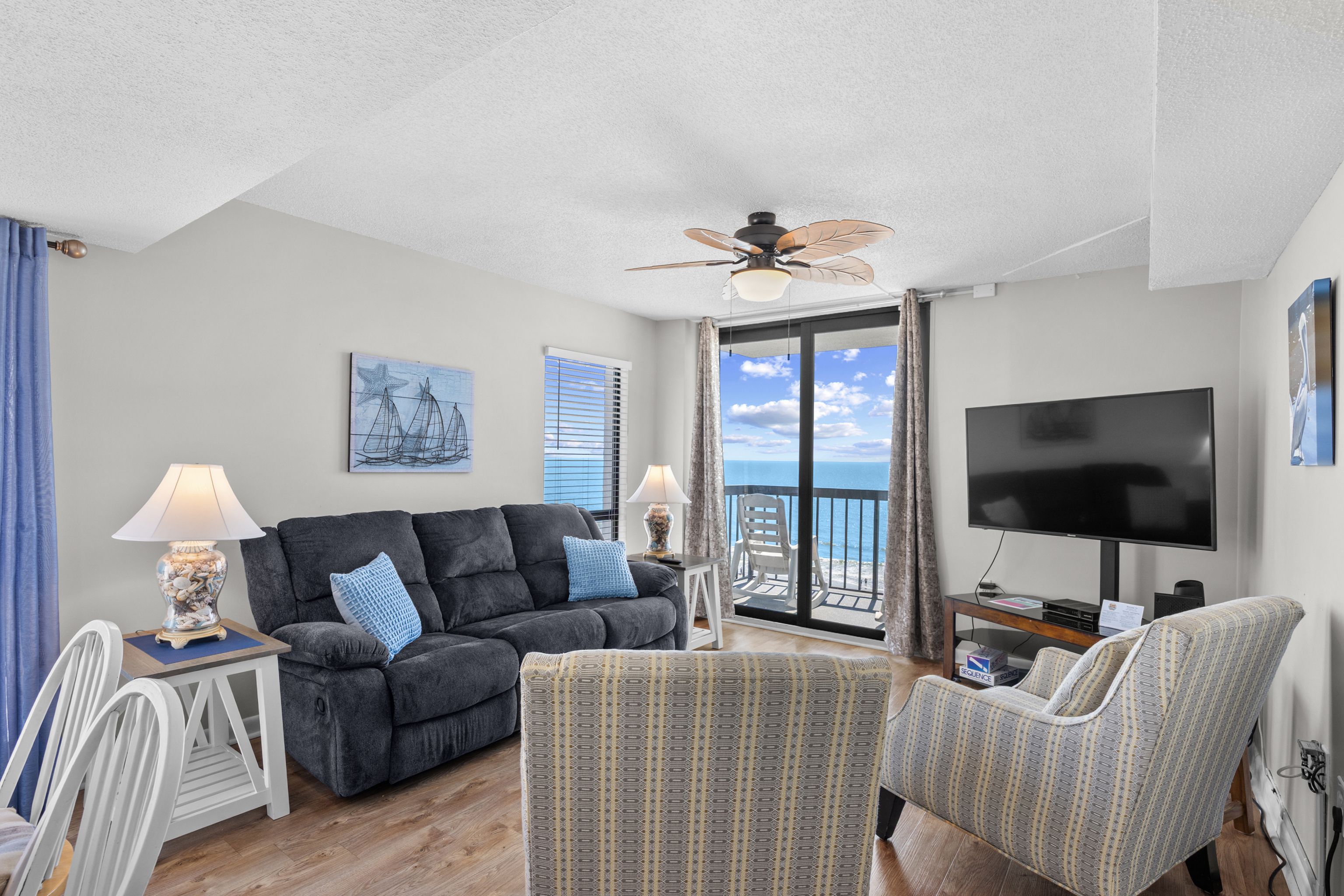 A living room with a sofa, armchairs, a TV, a ceiling fan, and a view of the ocean.