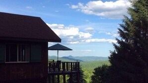 A wooden cabin with a deck and a large umbrella, overlooking a forested landscape with distant mountains.