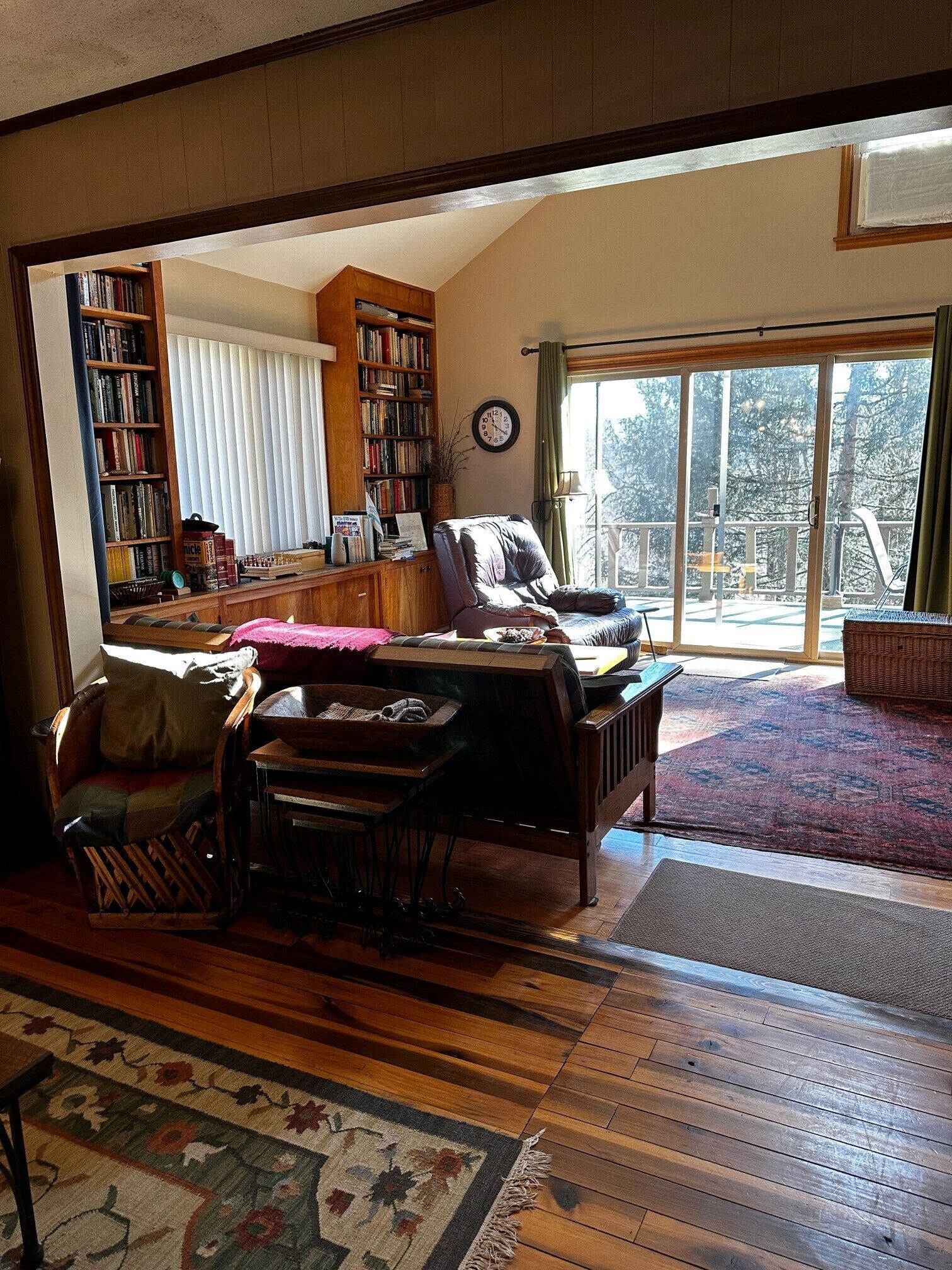 A cozy living room with a wooden floor, a large bookshelf, a comfortable sofa, and a view of a balcony with a railing.
